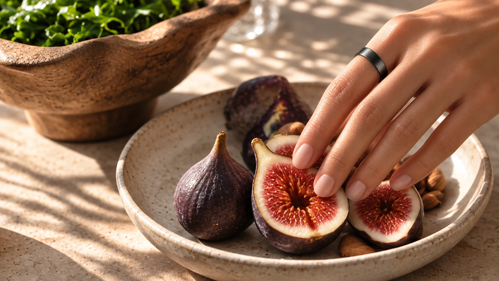 A hand wearing a dark smart ring reaching toward fresh cut figs and a bowl of salad